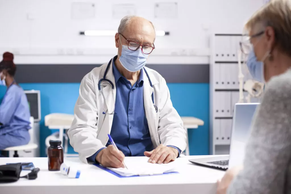 Elderly medic specialist in lab coat consults older woman, both masked, reviewing health details on clipboard. Retired male physician taking notes at medical appointment of female pensioner patient.